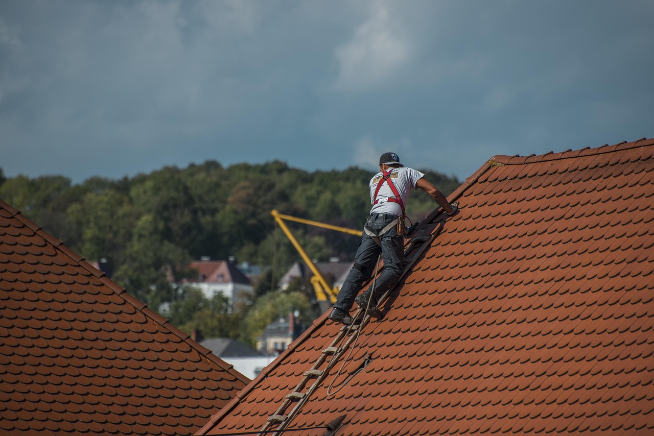 Roofing contractor wearing safety harness working on a steep red tile roof, inspecting damage for an insurance claim.